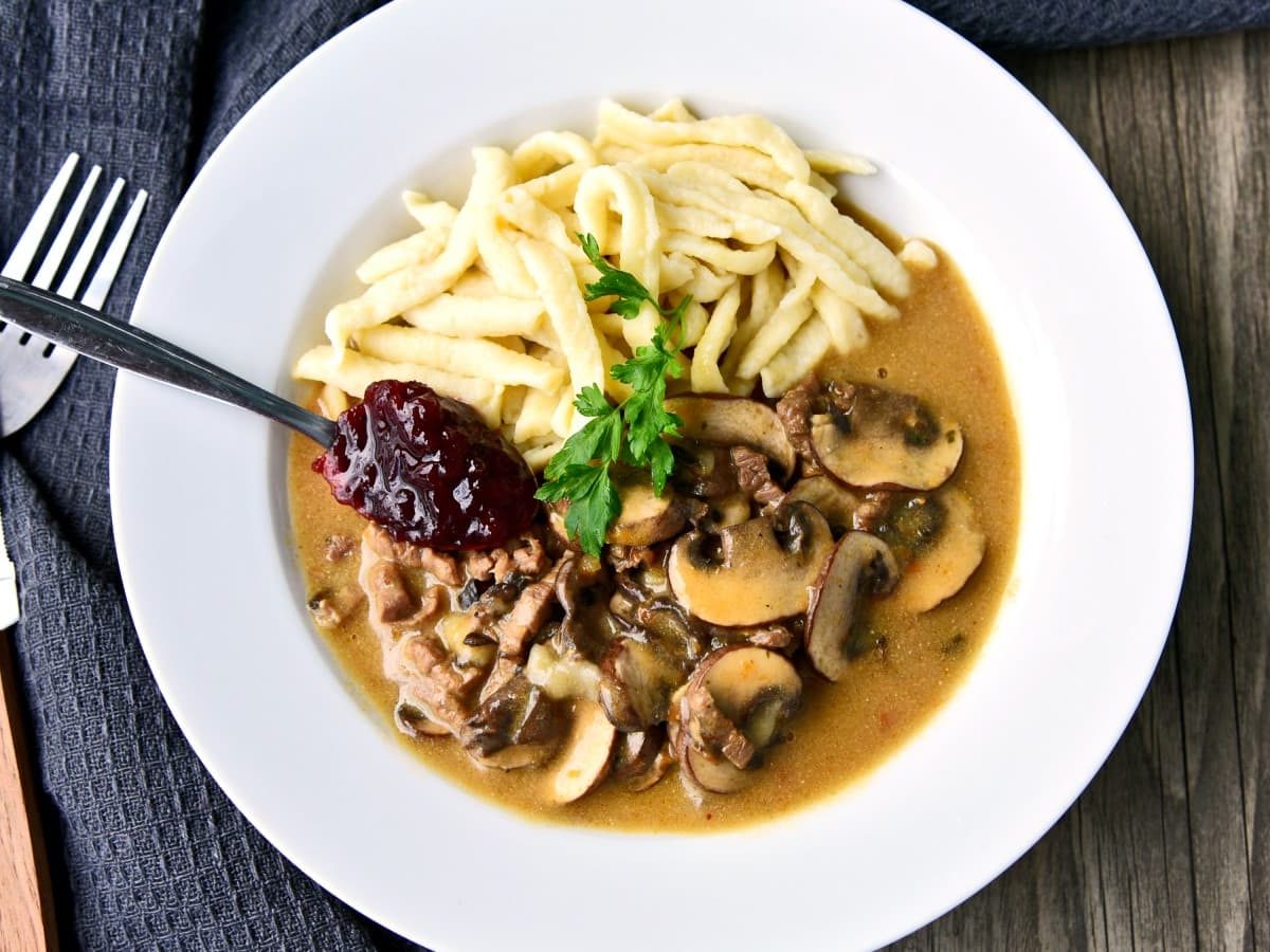 Ein veganes Champignon-Rahm-Geschnetzeltes mit Spätzle in einem weißen Teller vor einem hölzernen Hintergrund. Links davon ein schwarzes Geschirrtuch, darauf ein Messer und eine Gabel.