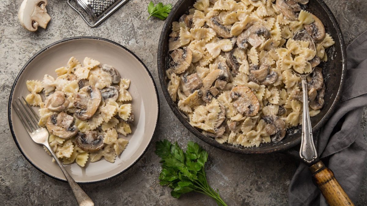 Ein Teller und eine Pfanne Walnuss-Pilz-Pasta mit Farfalle-Nudeln in der Draufsicht, daneben geschnittene Champignons und Kräuter.