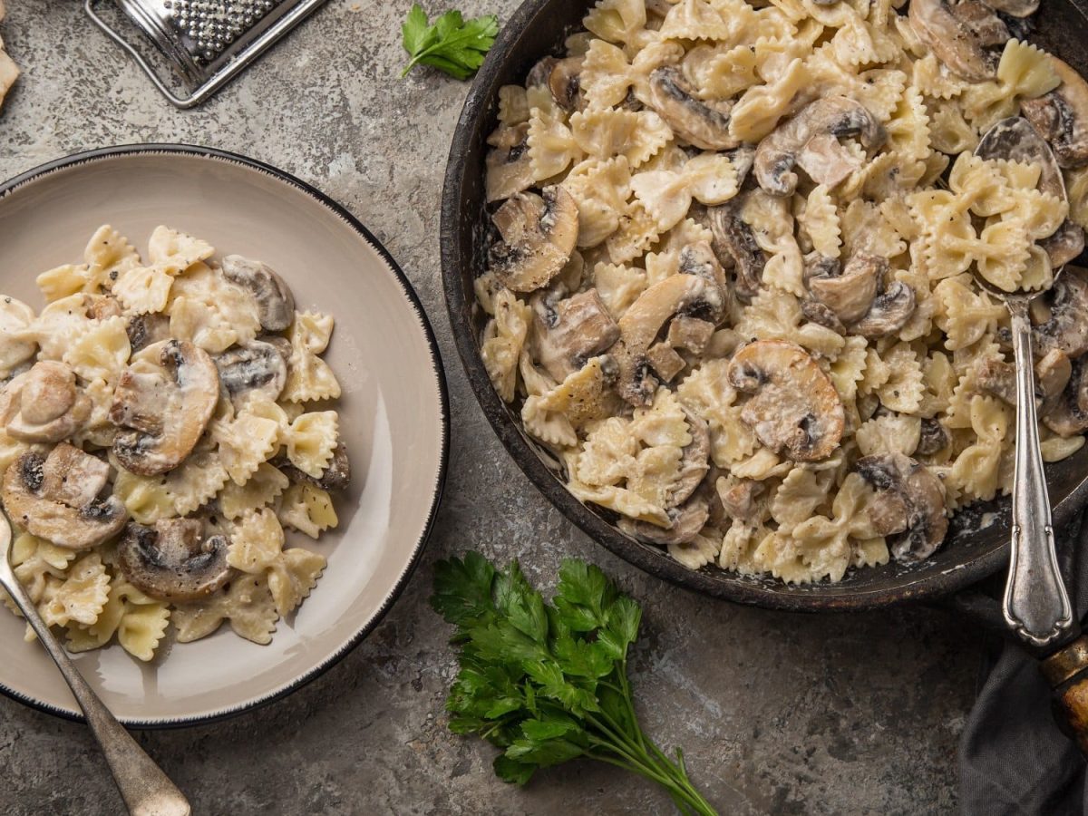 Ein Teller und eine Pfanne Walnuss-Pilz-Pasta mit Farfalle-Nudeln in der Draufsicht, daneben geschnittene Champignons und Kräuter.