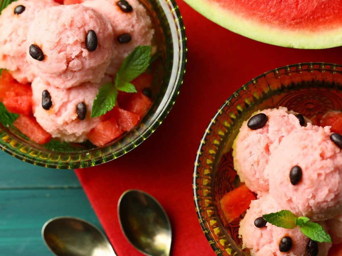 Draufsicht: Zwei Portionen selbst gemachtes Wassermelonen-Eis, eine angeschnittene Melone und zwei kleine Löffel.