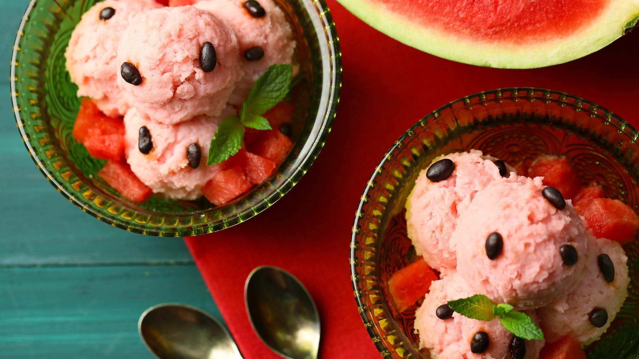 Draufsicht: Zwei Portionen selbst gemachtes Wassermelonen-Eis, eine angeschnittene Melone und zwei kleine Löffel.