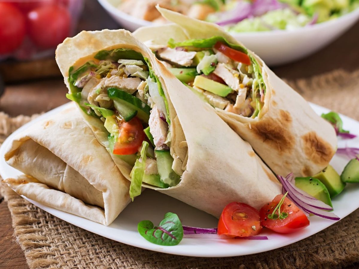 Ein weißer Teller mit dem Thunfisch-Wrap auf einem Holztisch. Im Hintergrund Salatzutaten und alles auf einem rustikalen Holztisch mit brauner Tisch-Matte seitlich fotografiert.