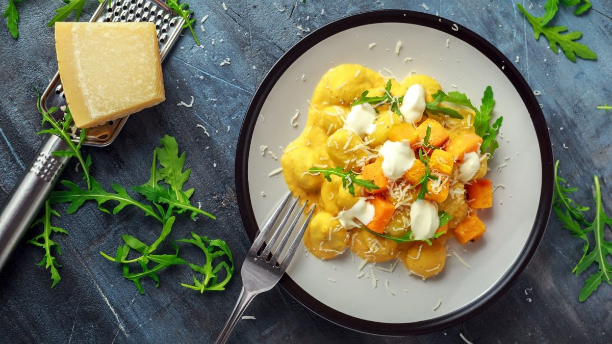 Ein weißer Teller mit Gnocchi in Kürbis-Ricotta-Creme neben einem Teller Rucola.