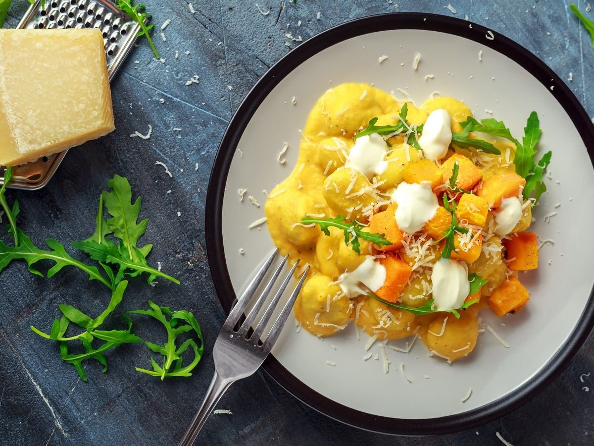 Ein weißer Teller mit Gnocchi in Kürbis-Ricotta-Creme neben einem Teller Rucola.