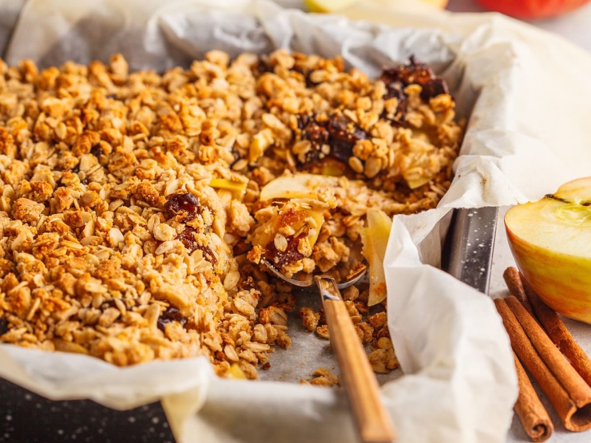 Ein Blech mit dem Apfel-Crumble-Cake auf einem hellen tisch mit Zimtstangen. Alles seitlich und nah fotografiert.