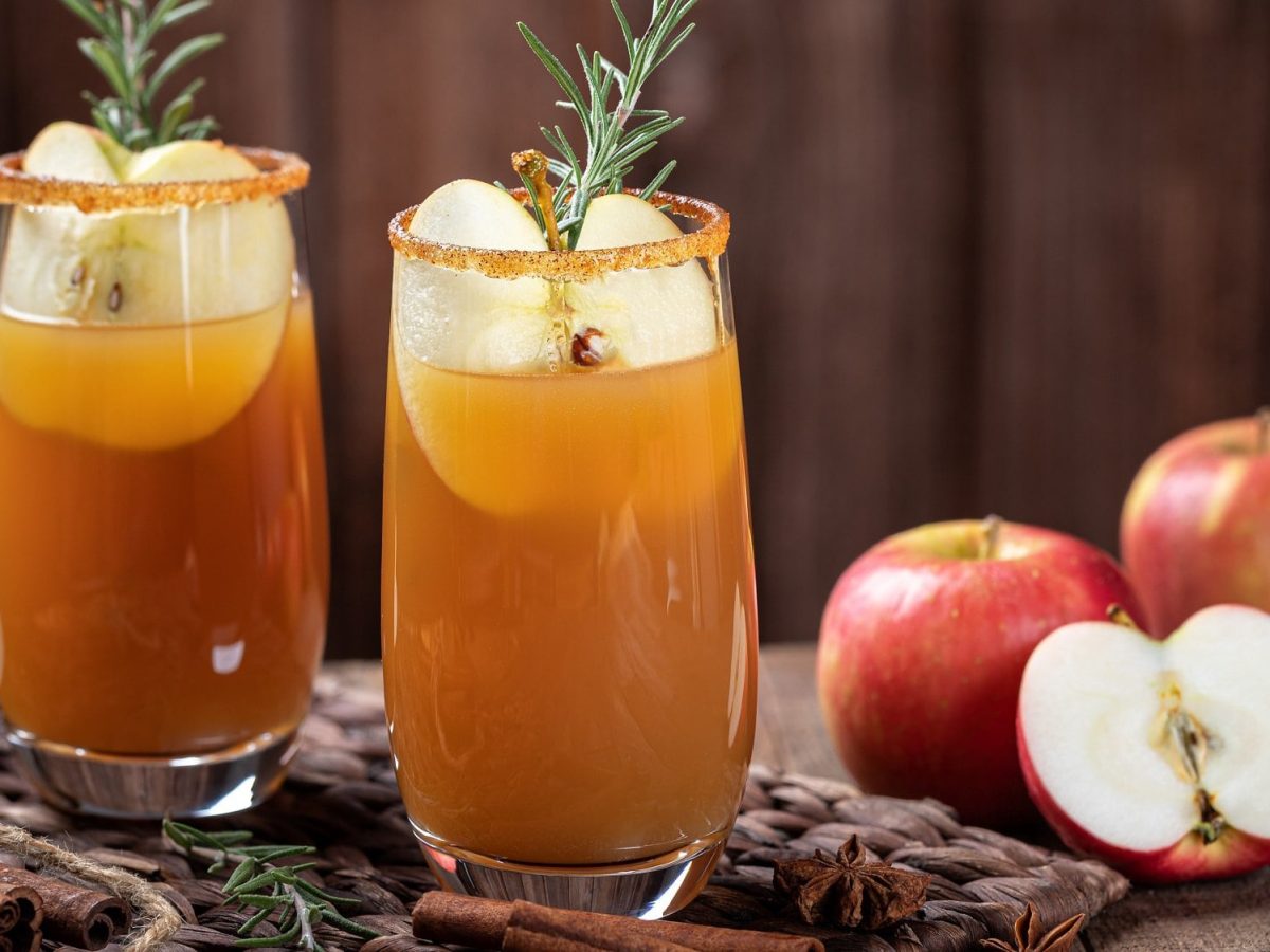 2 Gläser Autumn Spice Cocktail mit Eis, einem Stück Apfel und Rosmarin auf einem Brett, daneben ein angeschnittener Apfel und Zimtstangen.