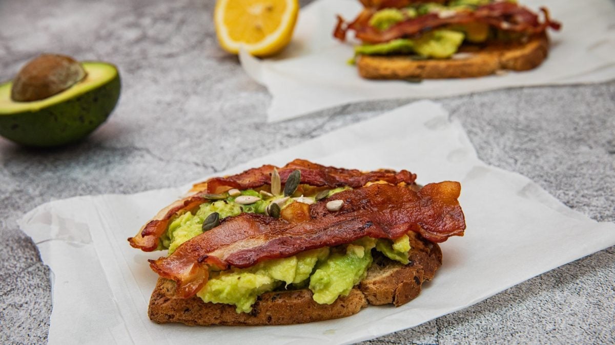 Schräge Draufsicht von oben: Heller Untergrund und zwei Bacon-Avocado-Stullen liegen auf weißem Papier. Im Hintergrund eine halbe Avocado mit Kern und eine halbe Zitrone.