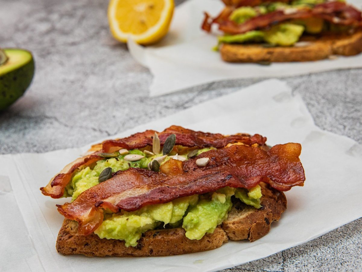 Schräge Draufsicht von oben: Heller Untergrund und zwei Bacon-Avocado-Stullen liegen auf weißem Papier. Im Hintergrund eine halbe Avocado mit Kern und eine halbe Zitrone.