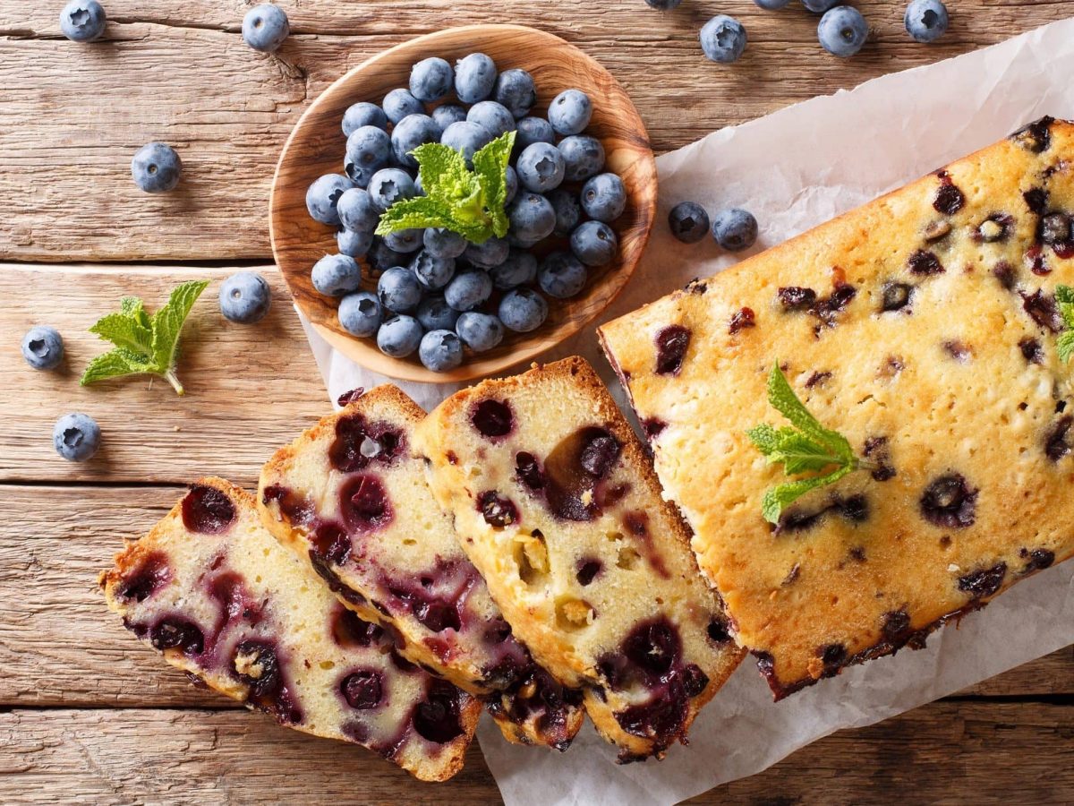 Draufsicht: Holzboden auf dem steht ein weißes Brettchen mit einem Heidelbeer-Zitronen-Kuchen. Daneben ist eine runde Schale mit Heidelbeeren.