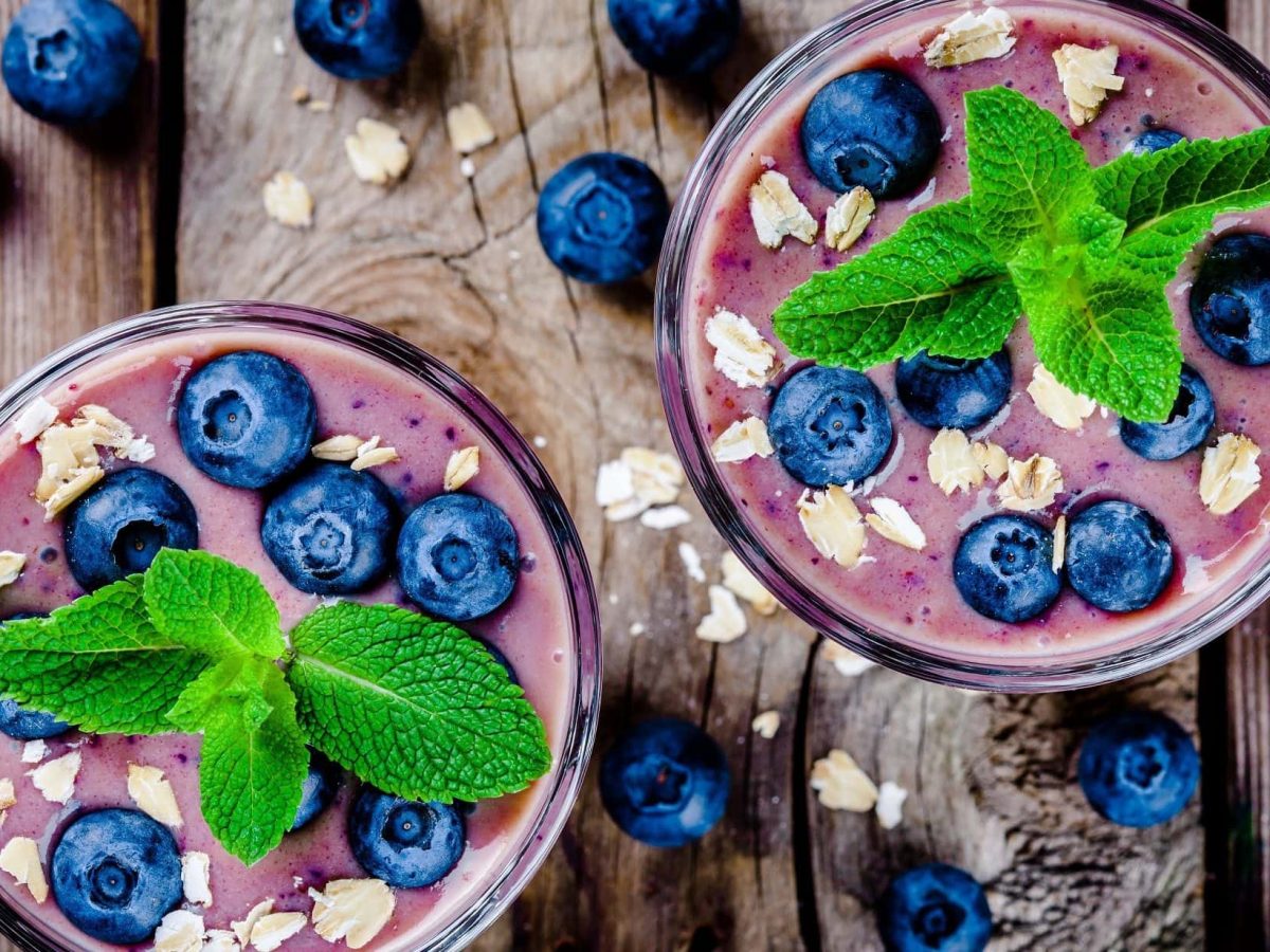 Draufsicht: Untergrund aus Holz, zwei Gläser Blaubeeren-Bananen-Smoothie mit Haferflocken und Minze. Drum herum liegen Blaubeeren.