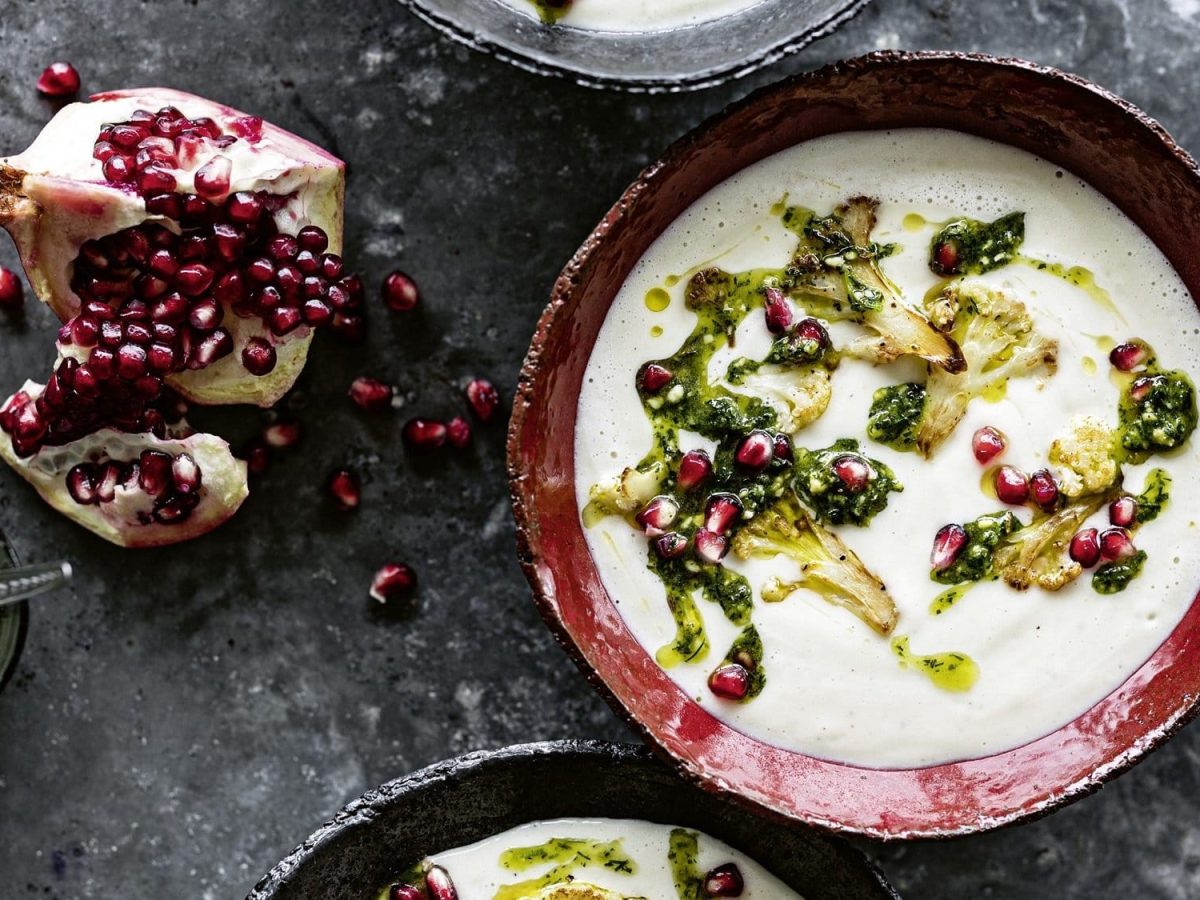 Eine Schüssel Blumenkohlsuppe mit Tahin und Granatapfelpesto, daneben ein geöffneter Granatapfel und zwei weitere Schüsseln, die man nur angeschnitten sieht, Draufsicht.