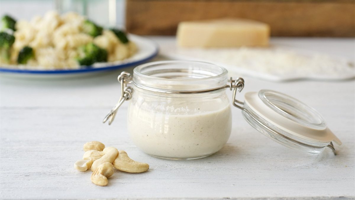 Ein offenes Weckglas mit Cashew-Cremesauce, daneben liegen ein paar Cashewkerne, im Hintergrund unscharf ein Teller Nudeln mit Brokkoli und Kücheneschirr.