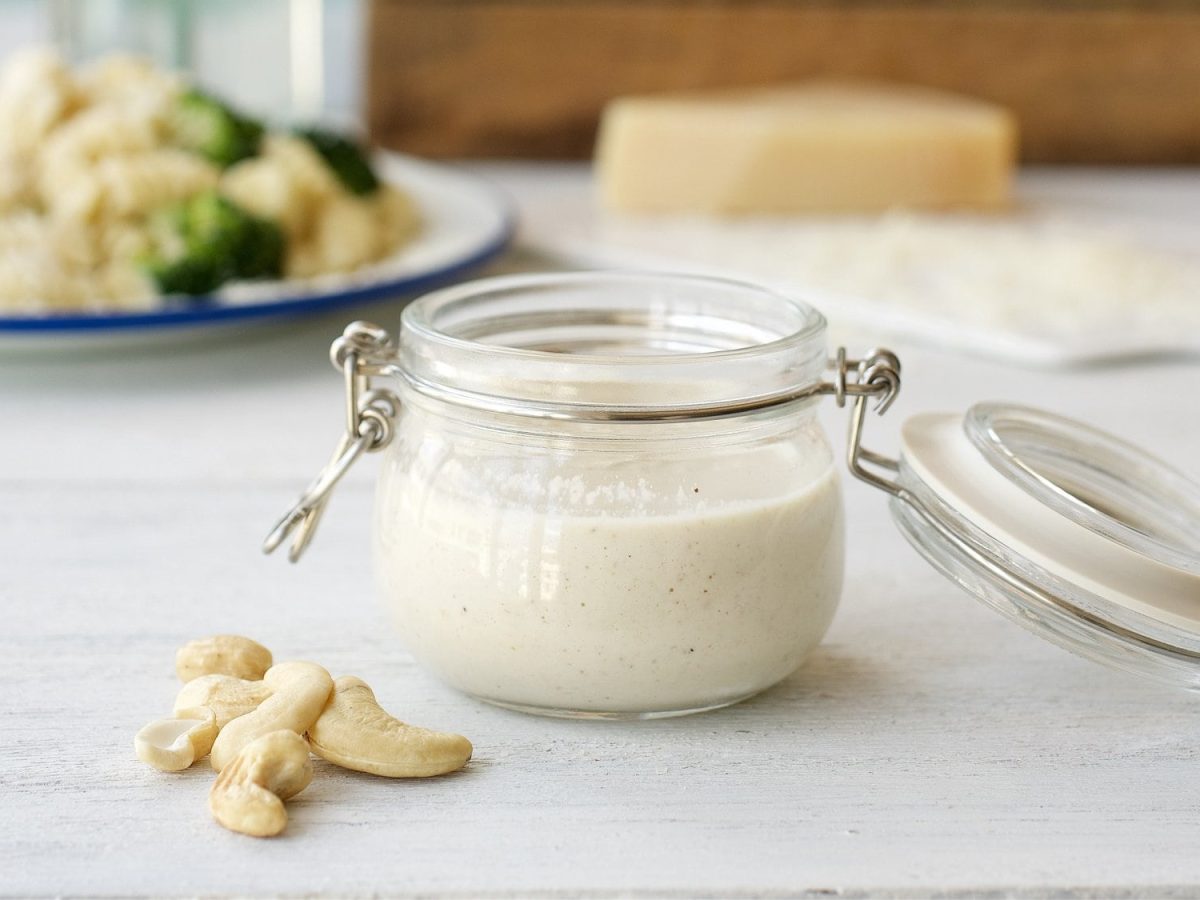 Ein offenes Weckglas mit Cashew-Cremesauce, daneben liegen ein paar Cashewkerne, im Hintergrund unscharf ein Teller Nudeln mit Brokkoli und Kücheneschirr.