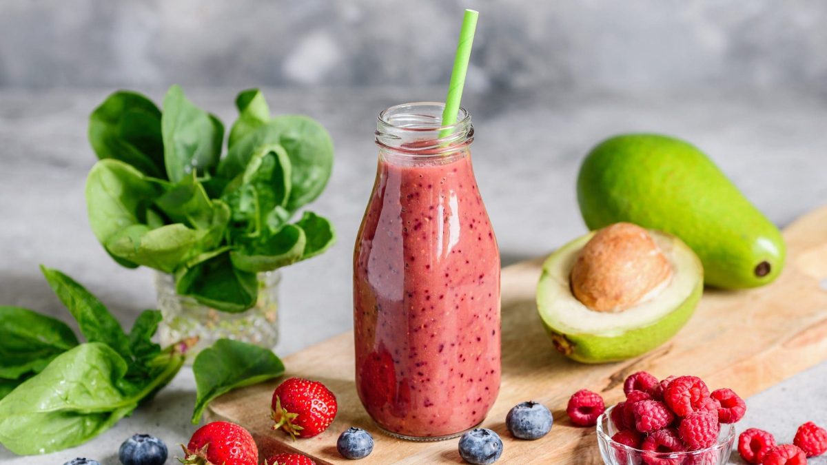 Frontal: Ein roter Avocado-Beeren-Kefir in einer Flasche mit einem Strohhalm drin. Drumherum liegen frische Erdbeeren und Blaubeeren sowie eine ganze und eine halbe Avocado.