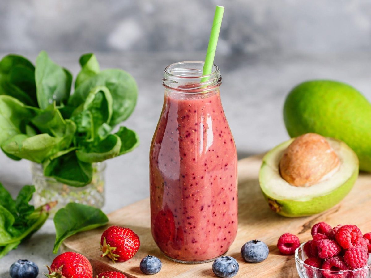 Frontal: Ein roter Avocado-Beeren-Kefir in einer Flasche mit einem Strohhalm drin. Drumherum liegen frische Erdbeeren und Blaubeeren sowie eine ganze und eine halbe Avocado.