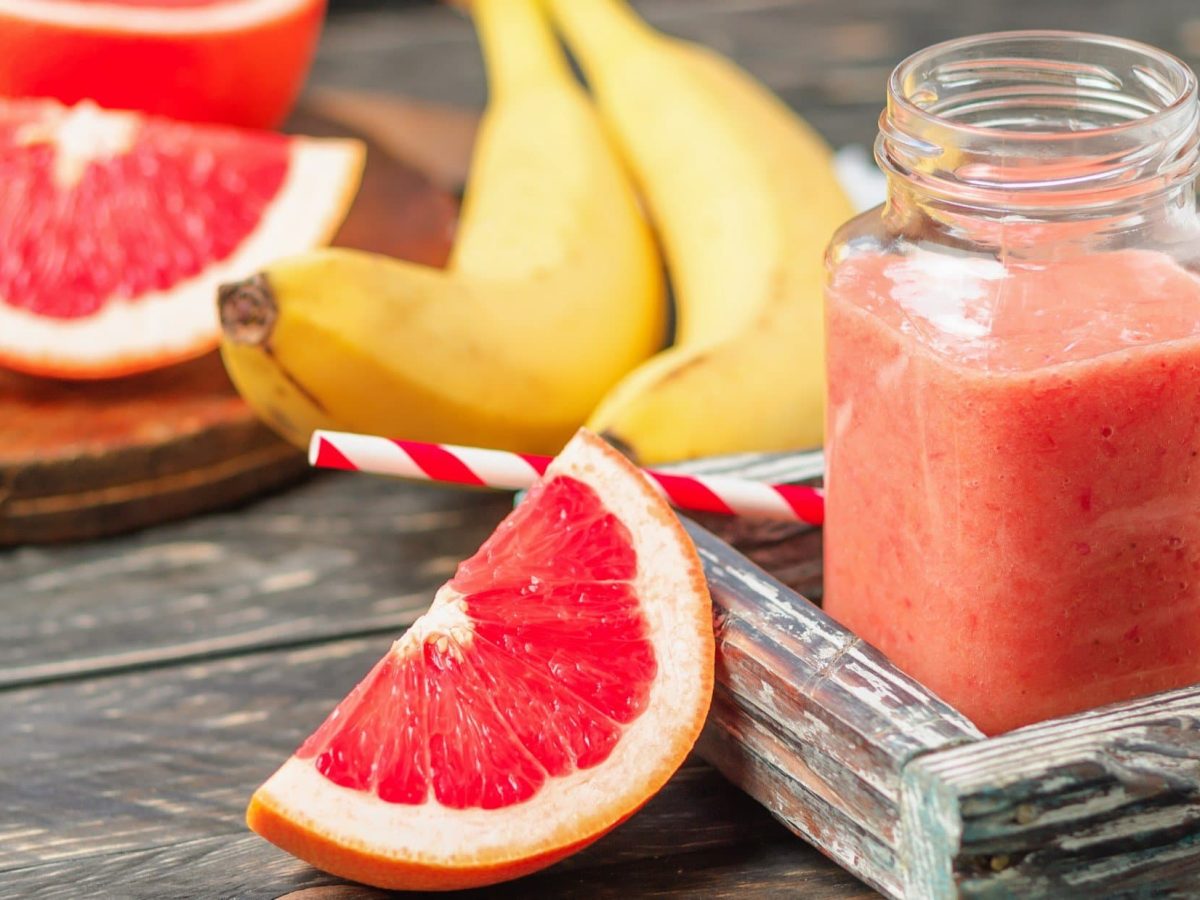 Frontal: Ein großes Glas mit einem Grapefruit-Bananen-Smoothie. Drumherum liegen Bananen und Grapefruits.