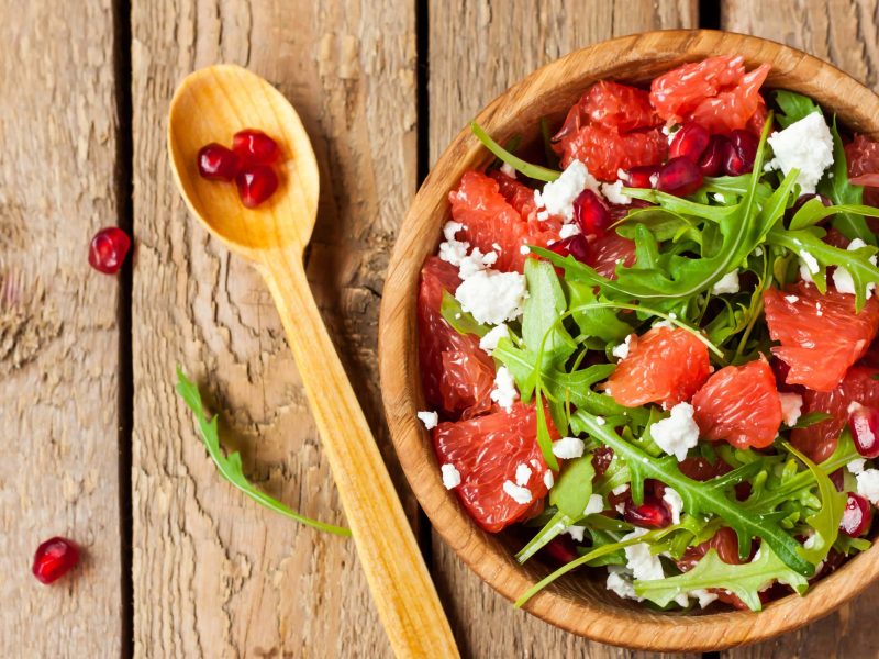 Draufsicht: Eine Schüssel Grapefruit-Salat mit Feta und Granatapfelkernen auf einem Untergrund aus Holz. Daneben liegt ein Löffel aus Holz.