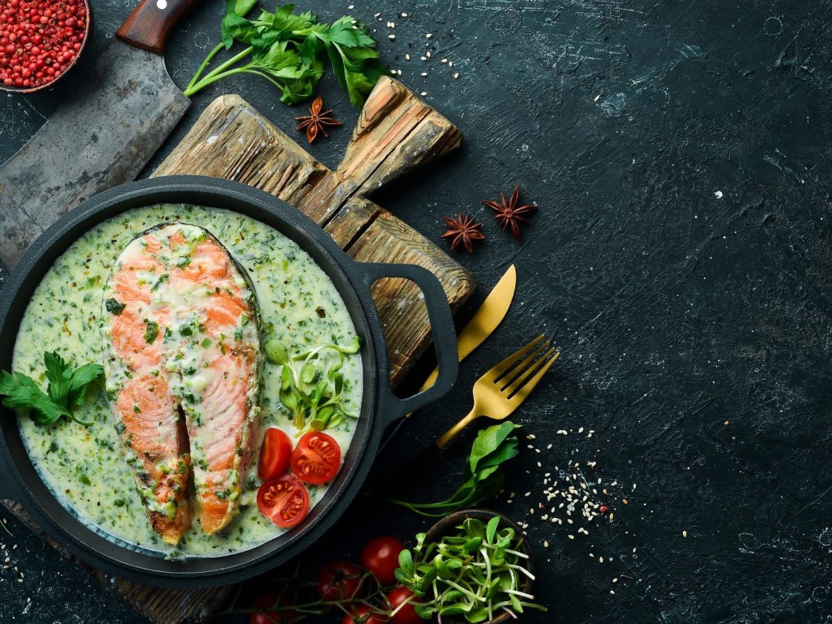 Draufsicht: Auf dunklem Untergrund steht eine Pfanne mit Lachs in Spinat-Sahnesauce. Drum herum liegen Kräuter.