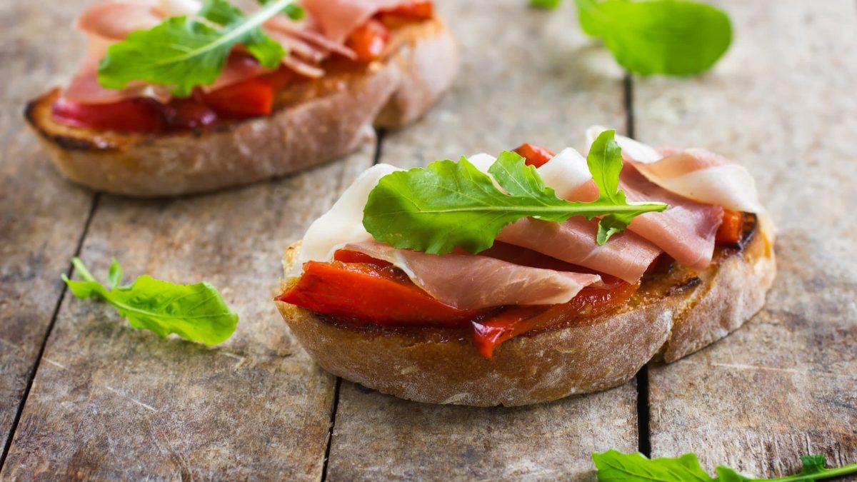 Draufsicht schräg: zwei Schinken-Bruschetta mit gerösteter Paprika auf einem Untergrund aus Holz.