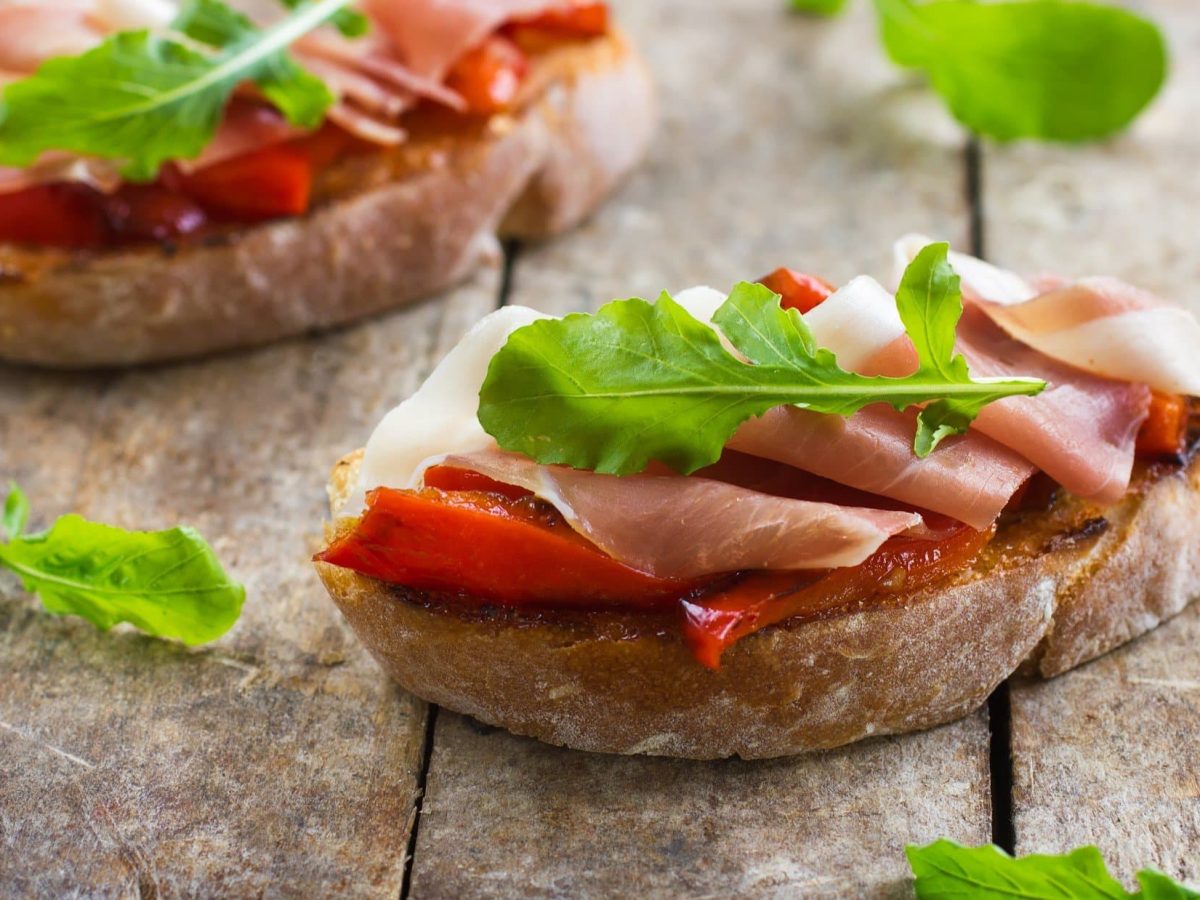 Draufsicht schräg: zwei Schinken-Bruschetta mit gerösteter Paprika auf einem Untergrund aus Holz.