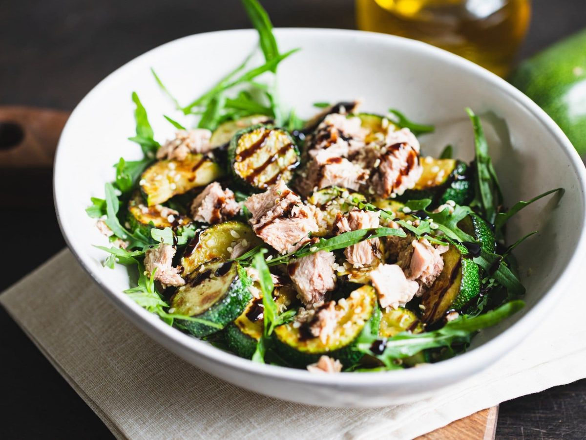 Draufsicht: eine weiße Schüssel Thunfisch-Salat mit gegrillter Zucchini auf einem Tuch, das auf einem Holzbrettchen liegt. Im Hintergrund ein Teil einer Zucchini und eine Flasche Öl.