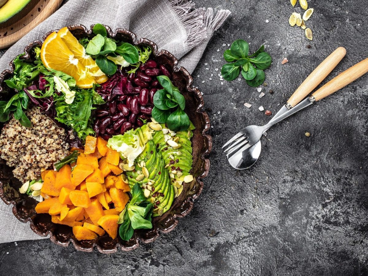 Fitness-Bowl mit Avocado, Feldsalat, Quinoa, Süßkartoffel, roten Bohnen und Orangenscheiben in der Draufsicht, daneben Besteck und ein paar Zutaten.