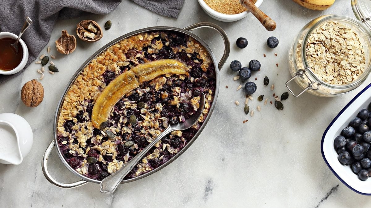 Gebackene Blaubeer-Bananen-Haferflocken in einer Auflaufform in der Draufsicht, daneben frische Beeren, eine Schüssel Haferflocken, Walnüsse und mehr Zutaten.