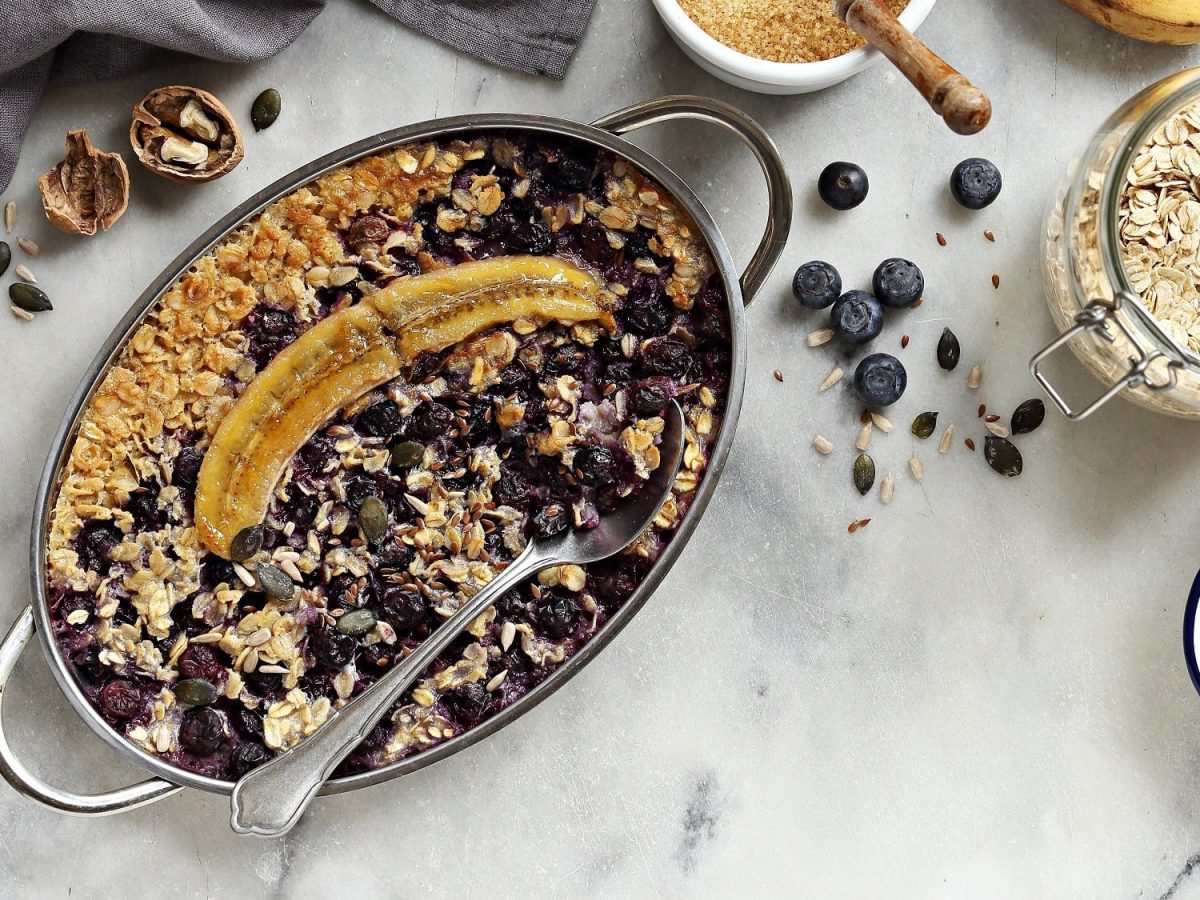 Gebackene Blaubeer-Bananen-Haferflocken in einer Auflaufform in der Draufsicht, daneben frische Beeren, eine Schüssel Haferflocken, Walnüsse und mehr Zutaten.