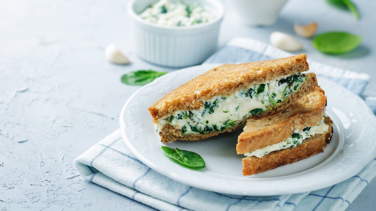 2 gegrillte Käse-Spinat-Sandwiches auf einem Teller, der auf einem karierten Handtuch steht, drumherum Knoblauchzehen und eine Schale mit der Füllung.