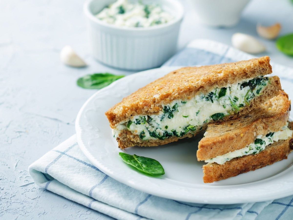 2 gegrillte Käse-Spinat-Sandwiches auf einem Teller, der auf einem karierten Handtuch steht, drumherum Knoblauchzehen und eine Schale mit der Füllung.