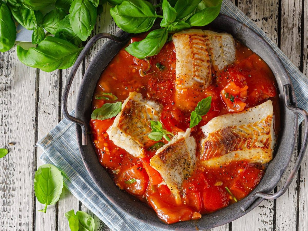 Draufsicht: auf einem hellen Holztisch steht eine Pfanne geschmortes Fischfilet auf Tomatensauce. Daneben liegen viele frische Basilikumblätter.