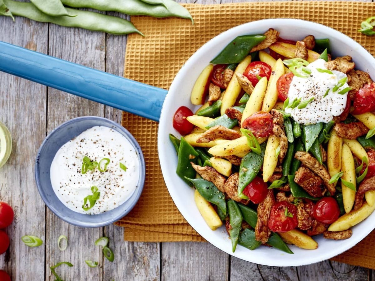 Eine Gyros-Schupfnudel-Pfanne auf einem gelben Geschirrtuch auf hölzernem Hintergrund. Links ein Schüsselchen mit Sour Cream, Tomaten und Öl. Oben auf dem Bild Bohnen.