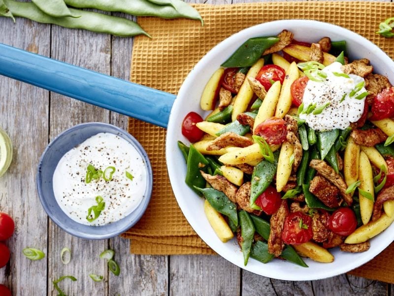 Eine Gyros-Schupfnudel-Pfanne auf einem gelben Geschirrtuch auf hölzernem Hintergrund. Links ein Schüsselchen mit Sour Cream, Tomaten und Öl. Oben auf dem Bild Bohnen.