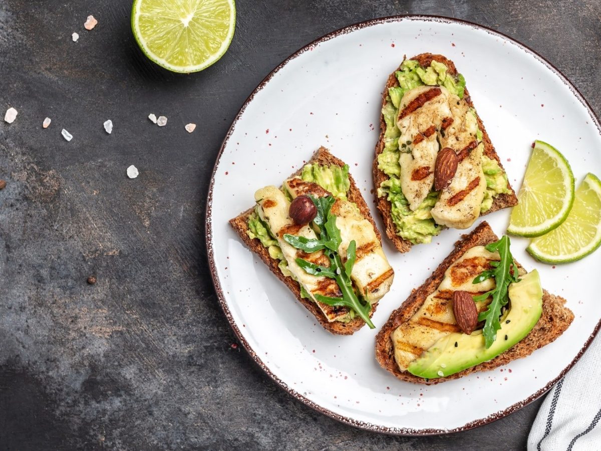 Halloumi-Avocado-Brot auf einem weißen Teller auf einem grauen Hintergrund. Oben eine halbe Zitrone, rechts ein weißes Geschirrtuch mit schwarzen Streifen und ein Pfefferstreuer.