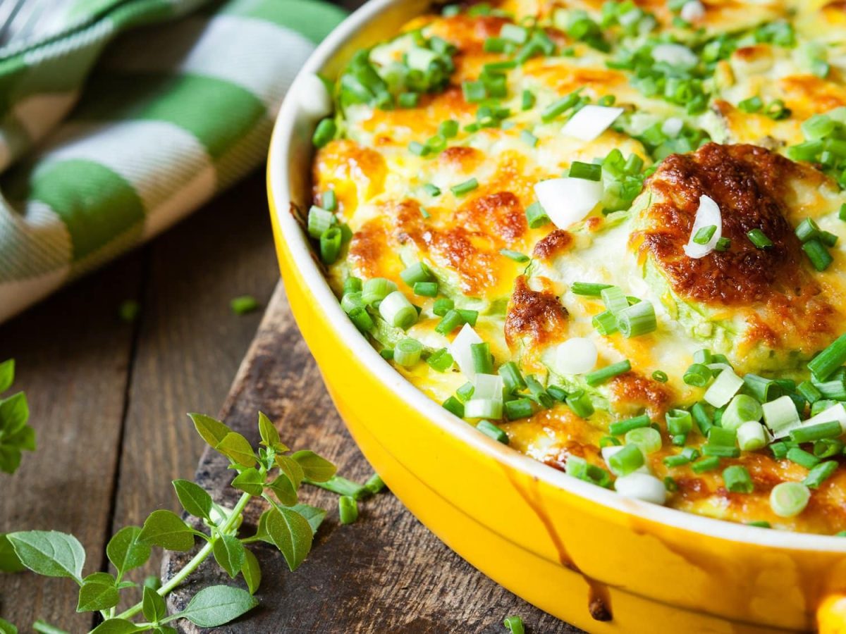 Herbstlicher Gemüseauflauf mit Käse überbacken und Frühlingszwiebeln bestreut, daneben Kräuter und ein grün-weiß kariertes Geschirrtuch.