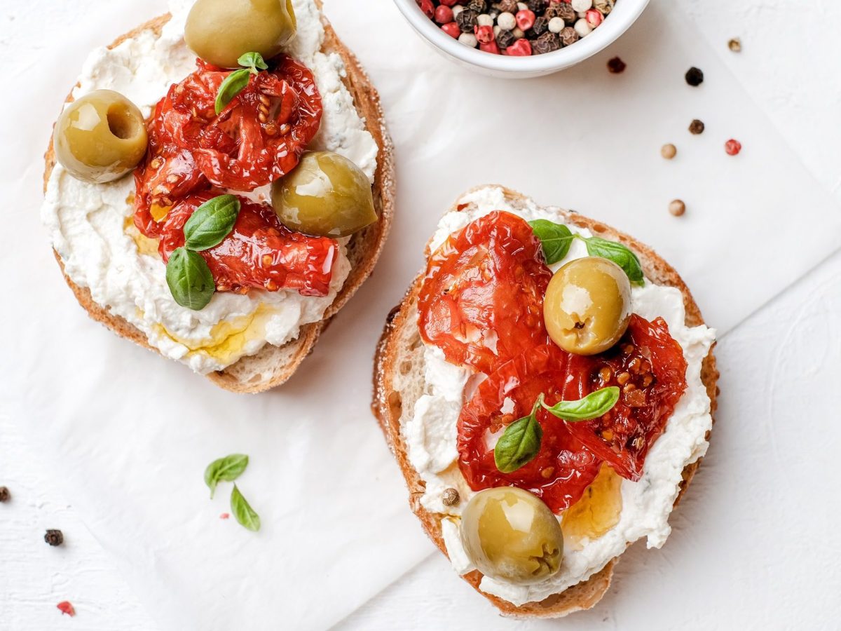 Draufsicht: Weißer Untergrund und zwei Käse-Bruschetta mit getrockneten Tomaten und Oliven.