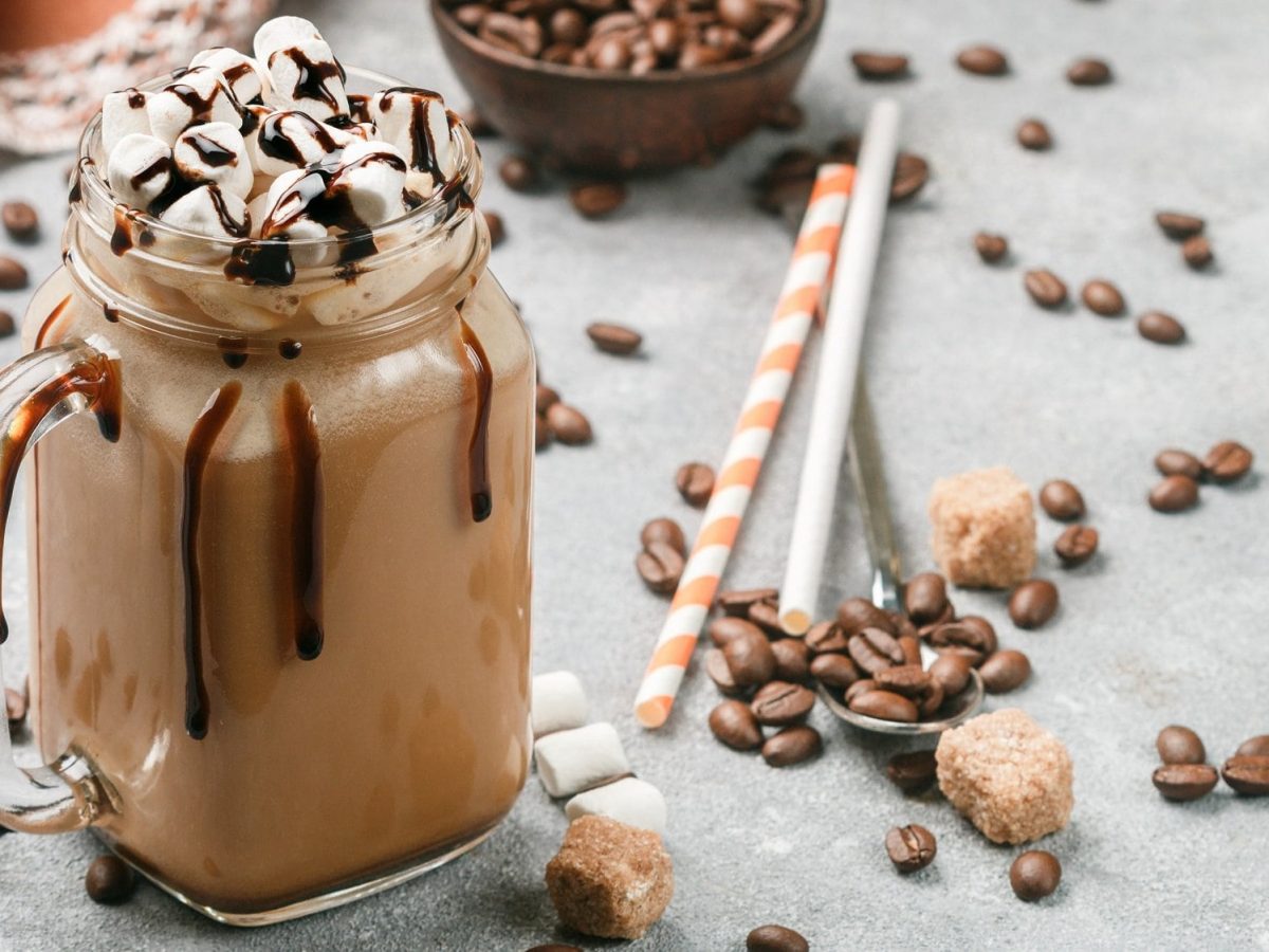 Kaffee-Shake in einem Glas mit Henkel, getoppft mit Marshmallows und Schokosauce, daneben Kaffeebohnen und Stohhalme.