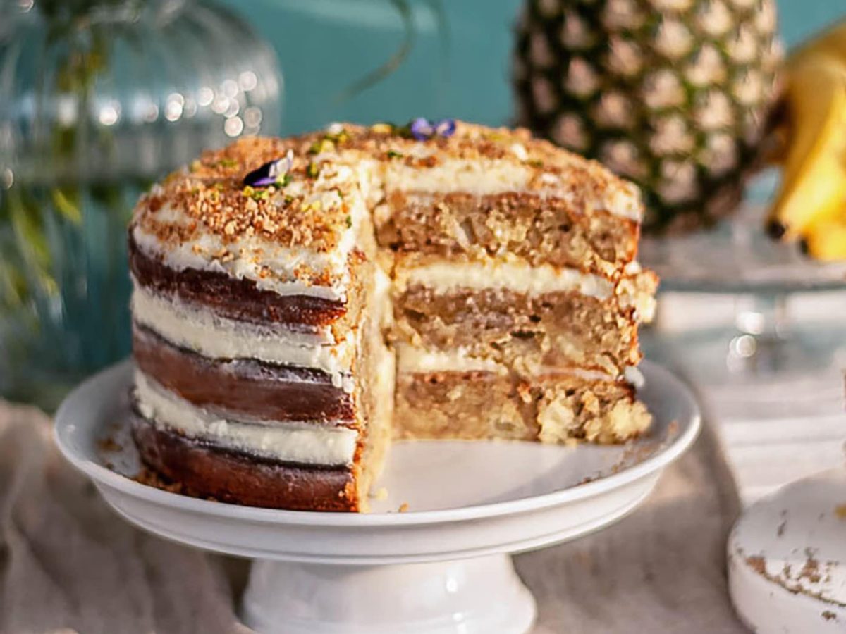 Ein Kolibri-Kuchen aus 3 Böden und Creme dazwischen und darauf auf einem Tortenständer, im Hintergrund eine Ananas, Bananen, eine Glasvase, daneben ein Teller mit einem Stück des Kuchens.