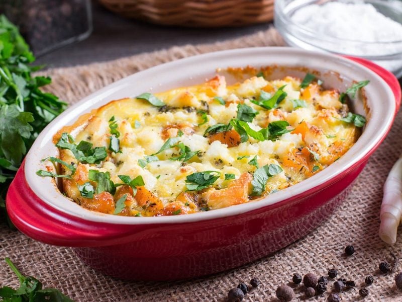 Ein Kürbis-Gnocchi-Auflauf in einer ovalen, roten Auflaufform, daneben Zutaten wie Frischkäse in einer Glasschale, Petersilie oder Lauchzwiebeln.