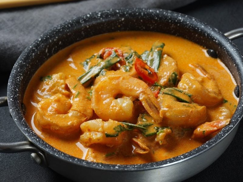 Eine Pfanne mit dem Curry auf einem grauen Tisch. Im dunklen Hintergrund sind unscharf Holz-Stäbchen zu erkennen. Alles von schräg oben fotografiert.