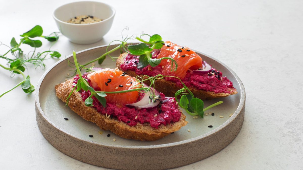 Draufsicht schräg: Heller Untergrung, grauer Teller mit zwei Lachs-Broten mit Rote Bete-Frischkäse und Erbsensprossen.