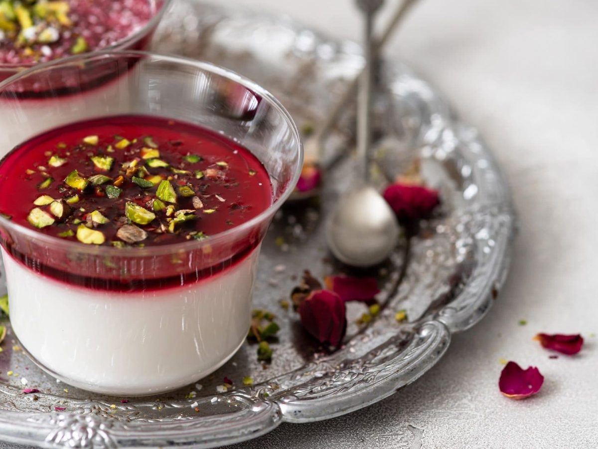 Draufsicht, schräg: Zwei Dessertgläser mit Malabi ,stehen auf einem orientalisch angehauchtem Tablett aus Metall. Drum herum liegen Rosenblüten.