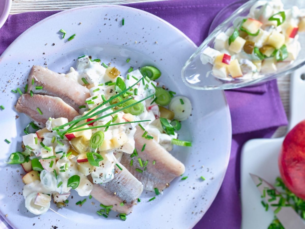 Matjes mit Apfel und Zwiebeln auf einem weißen Teller auf einem violetten Geschirrtuch. Rechts eine Sau­ci­e­re mit Joghurt-Mix, rechts unten ein Schneidebrett mit einem Apfel.