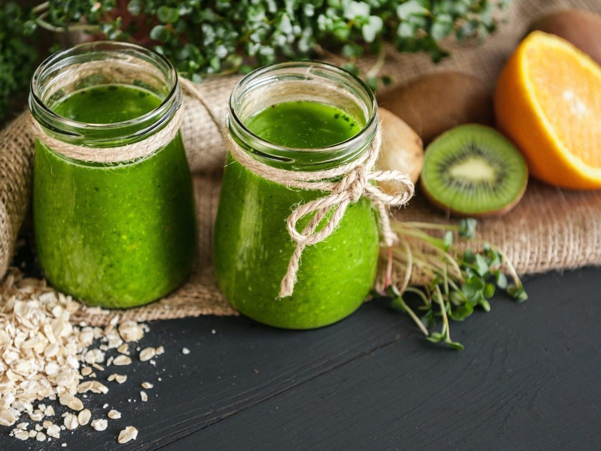 Zwei Gläser Microgreen-Smoothies auf einem dunklem Untergrund. Drum herum liegen Sprossen, Orangen und Kiwis.