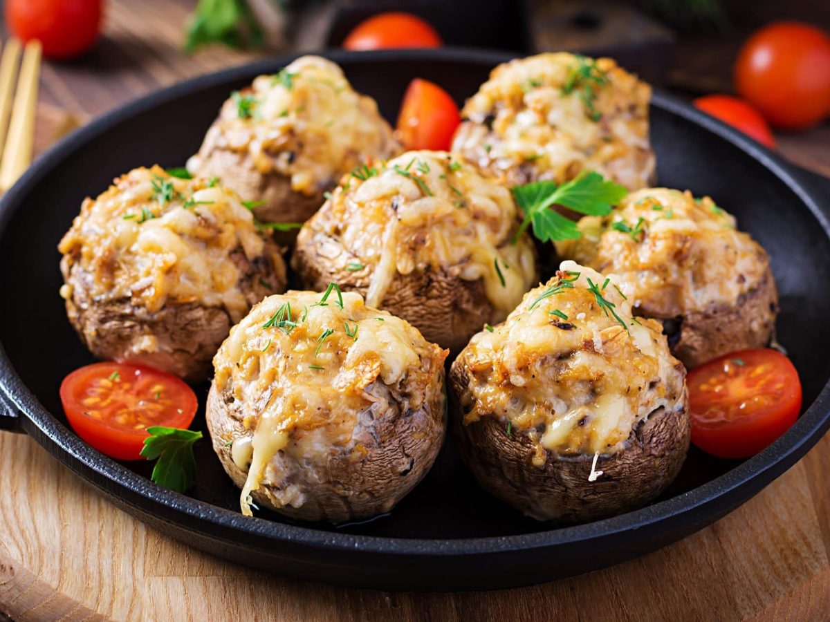 Draufsicht, schräg: eine gusseiserne Pfanne mit Ofen-Champignons mit Knofi-Hack-Füllung auf einem Holzbrett. Daneben liegt eine Gabel.