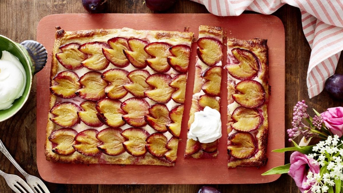 Eine Pflaumen-Blätterteig-Tarte auf einem roten Schneidebrett. Links davon eine Schüssel mit Schlagsahne und zwei Dessertgabeln. Rechts ein Geschirrtuch und Blumen. Oben auf dem Bild Pflaumen. Der Hintergrund ist hölzern.