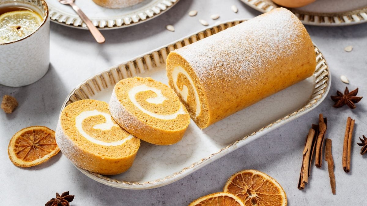 Eine weiße längliche Schale mit der Pumpkin Roll auf einem hellen, herbstlich dekoriertem Tisch mit Orangen, Zimt, Kürbis und Sternanis. Alles von schräg oben fotografiert.