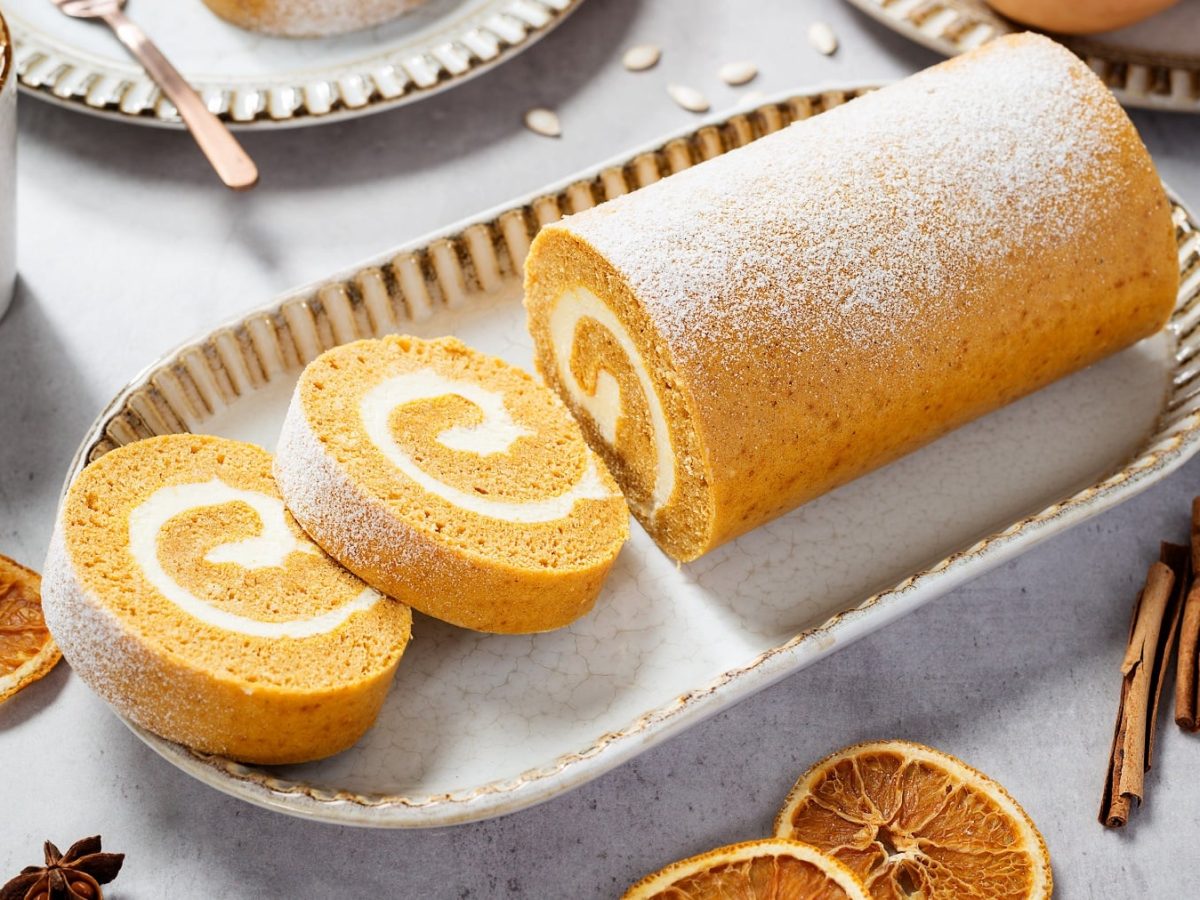 Eine weiße längliche Schale mit der Pumpkin Roll auf einem hellen, herbstlich dekoriertem Tisch mit Orangen, Zimt, Kürbis und Sternanis. Alles von schräg oben fotografiert.