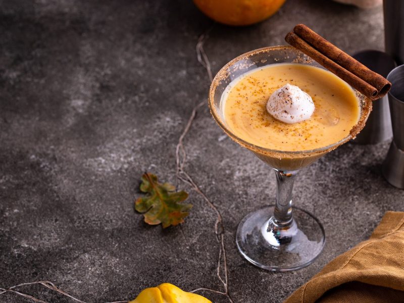 Ein Martiniglas mit Pumpkin Spice Likör und einem Klecks Sahne, garniert mit Zimtstangen, auf dunklem Untergrund, daneben ein Kürbis.