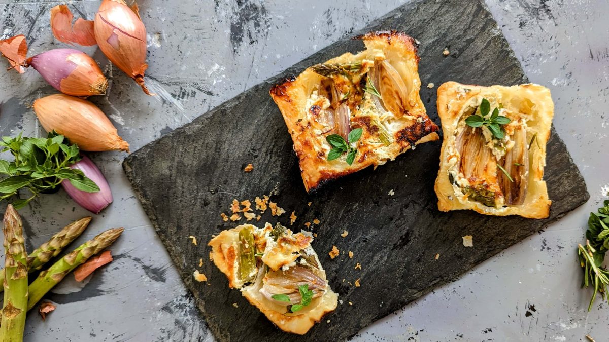 Eine Schieferplatte mit ein paar Stücken der Upside-Down-Tarte direkt von oben fotografiert. Alles auf einem Steintisch. Daneben noch grüner Spargel und Schalotten.